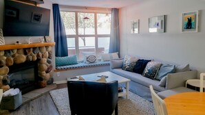 Een gezellige woonkamer met een stenen open haard, een bank met opvallende kussens, een bijzettafeltje en een aan de muur bevestigde televisie.