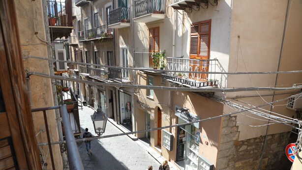 Une rue étroite, bordée de bâtiments des deux côtés, avec des balcons dotés de balustrades et une enseigne indiquant l’emplacement d’un magasin.