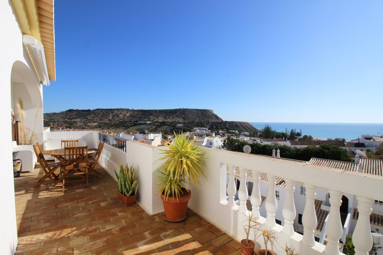 Un balcon avec une table et des chaises en bois, des plantes en pot et une vue sur la mer et les collines.