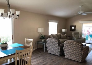 Uma sala de estar com sofá, duas poltronas, uma mesa de jantar com cadeiras, um lustre e um ventilador de teto.