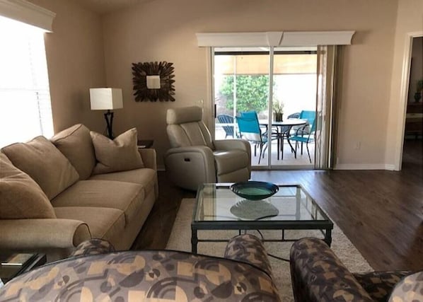 Una sala de estar con un sofá color beige, un sillón estampado, una mesa de centro de vidrio y una lámpara. Hay un ventanal corredero que da a un patio exterior con sillas y una mesa.