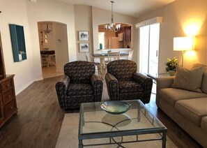 Una sala de estar con dos sillones estampados, una mesa de centro de vidrio, un sofá y una cocina al fondo.
