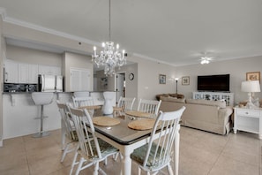 A dining area with a chandelier, white chairs, and a glass-top table. - Steps from the beach! Pool, Pickleball, and Tennis Courts! (Miramar Beach)