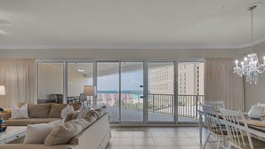 A spacious living room with a large sliding glass door leading to a balcony with a view of the sea and buildings. - Steps from the beach! Pool, Pickleball, and Tennis Courts! (Miramar Beach)