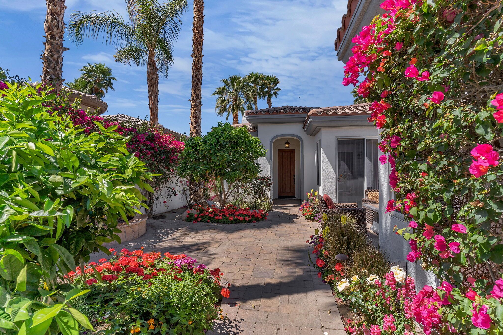 A pathway leading to a building with a white facade and a brown door, surrounded by lush greenery and vibrant flowers.