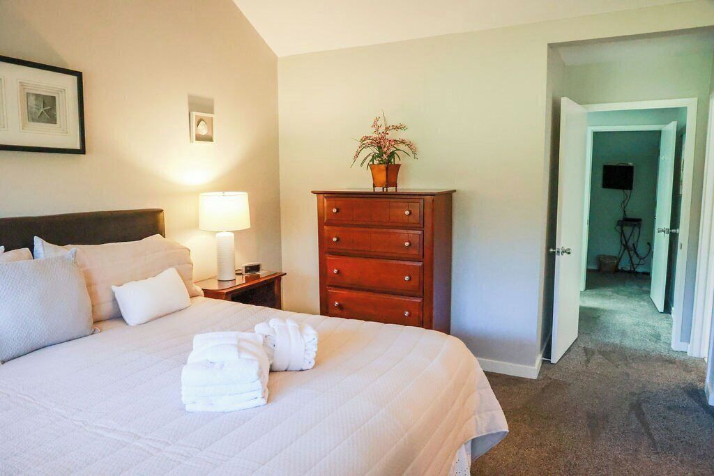Een net opgemaakt bed met witte lakens en handdoeken, een houten dressoir met een plant erop, en een lamp op een nachtkastje.