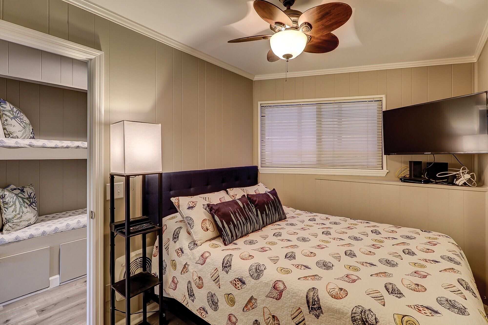 Une chambre à coucher comprenant un lit, un ventilateur de plafond, une fenêtre avec des stores, un téléviseur et des étagères intégrées.