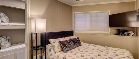 Une chambre à coucher comprenant un lit, un ventilateur de plafond, une fenêtre avec des stores, un téléviseur et des étagères intégrées.