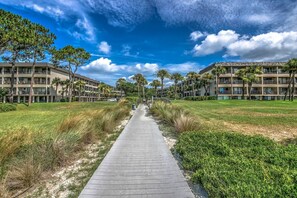 A pathway leading to a building with multiple floors and balconies, surrounded by greenery and palm trees.