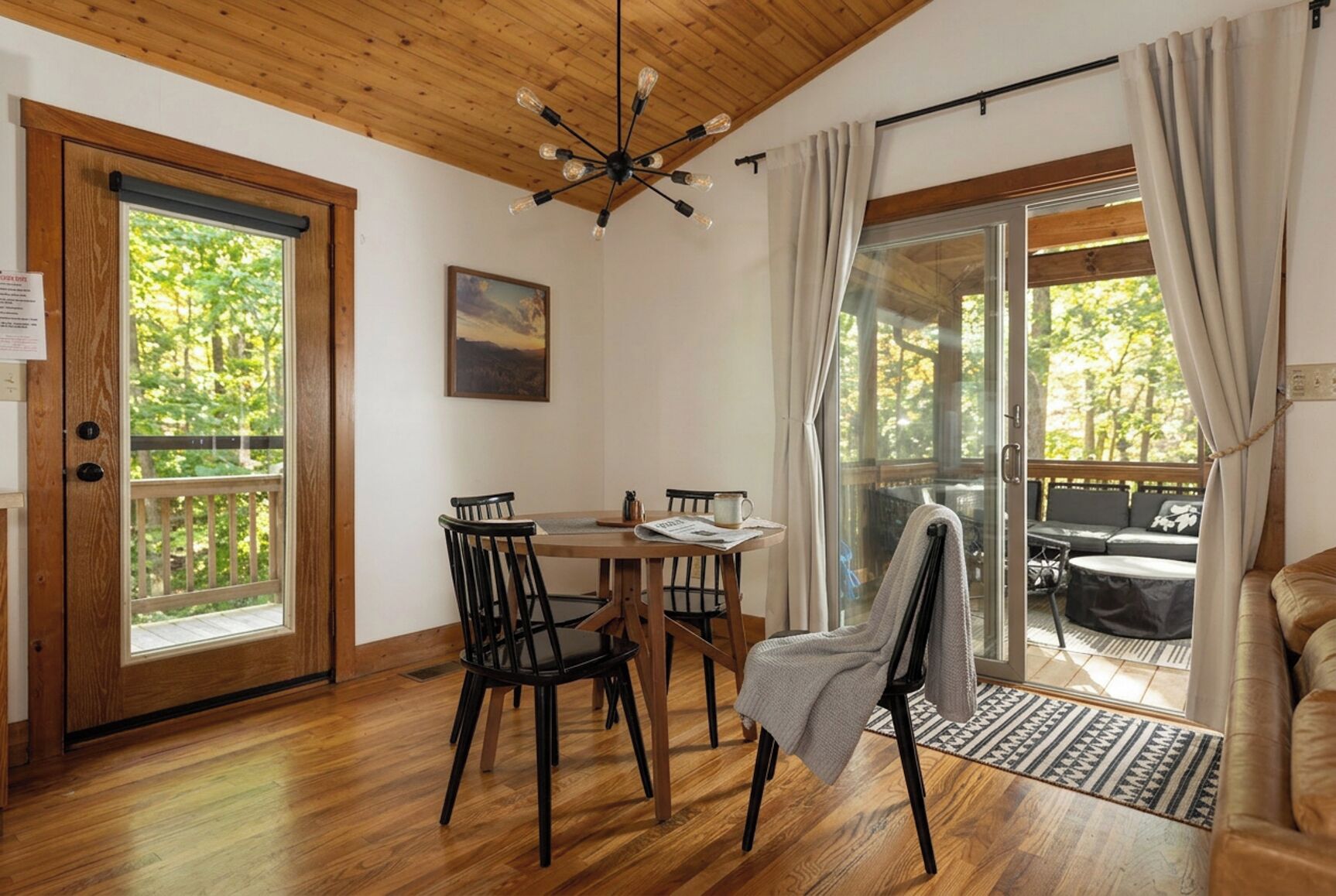 A wooden-floored room with a dining table, chairs, a sofa, and a large window with curtains.