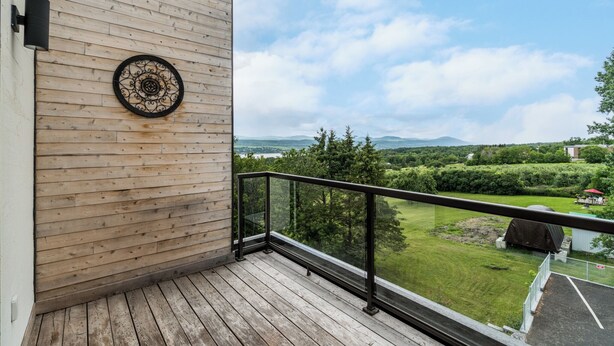 Un balcon en bois avec une balustrade en métal, une plaque décorative sur le mur et une vue sur un paysage verdoyant, avec des arbres et des collines à l’horizon.