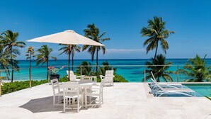 A terrace with white furniture, a large umbrella, and a view of the ocean and palm trees.