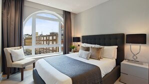 A modern hotel room with a large bed, a seating area, and a view of the cityscape through a large window.