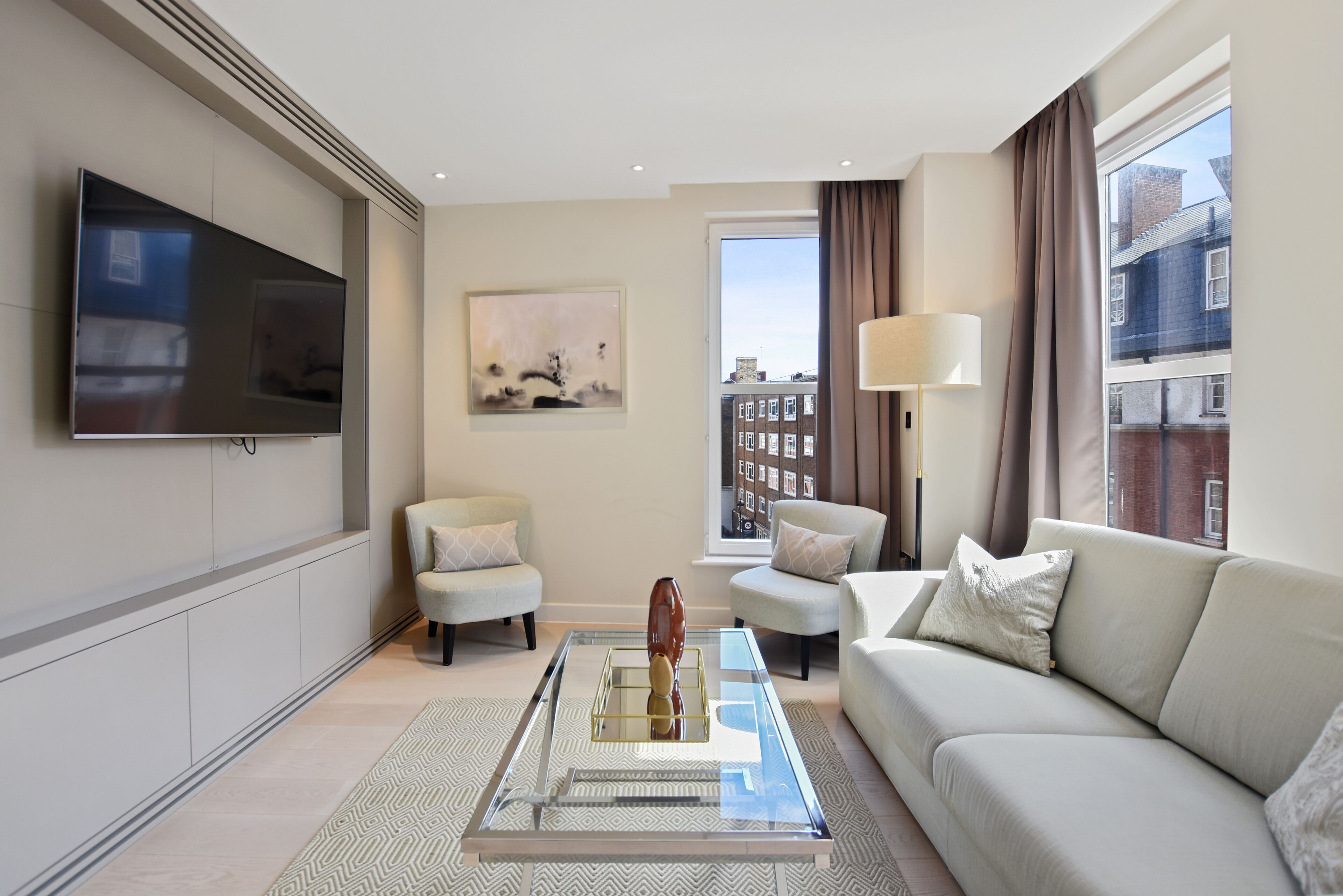 Una sala de estar moderna con un televisor de pantalla plana, dos sillones, un sofá, una mesa de centro de vidrio y un ventanal con cortinas.