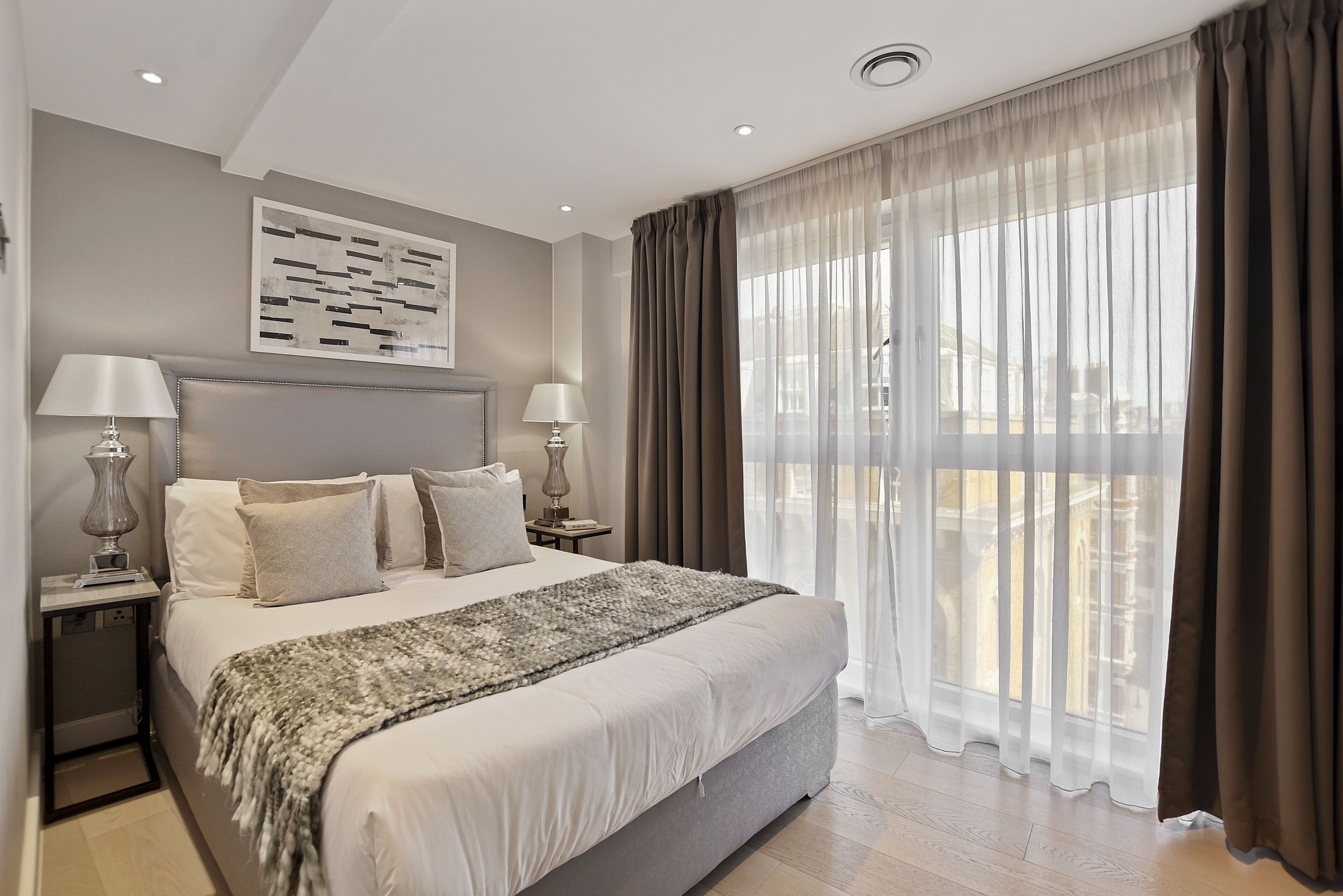 Een moderne slaapkamer met een groot bed, twee nachtkastjes met lampen, een decoratief wandobject en lichte gordijnen.