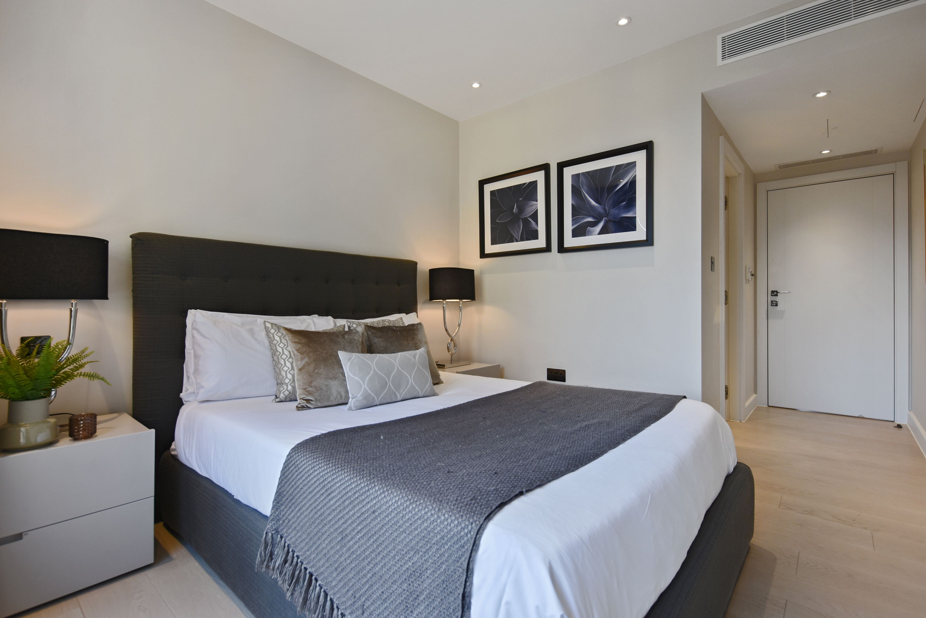 A modern bedroom with a large bed, two bedside tables, a nightstand, a potted plant, and two wall-mounted lamps.