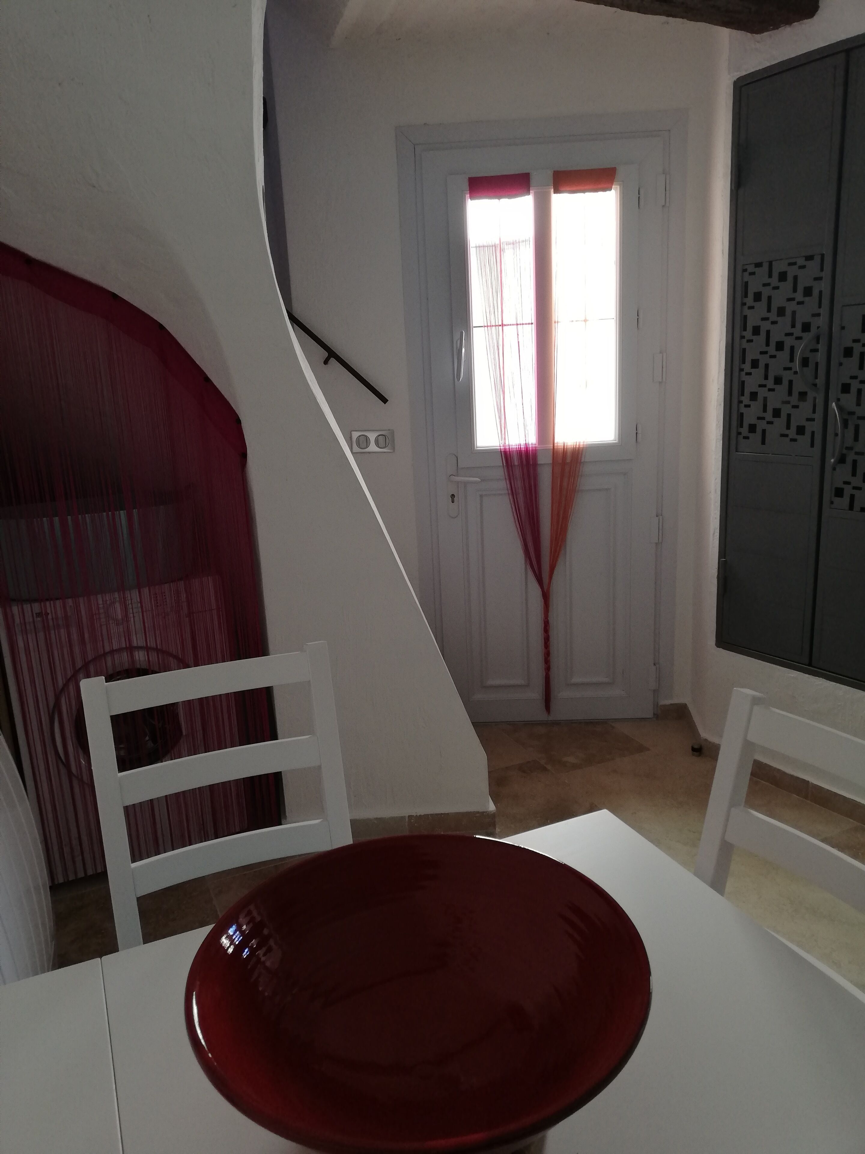 Un'area pranzo con un tavolo e sedie bianche, un centrotavola rosso e una finestra con tende.