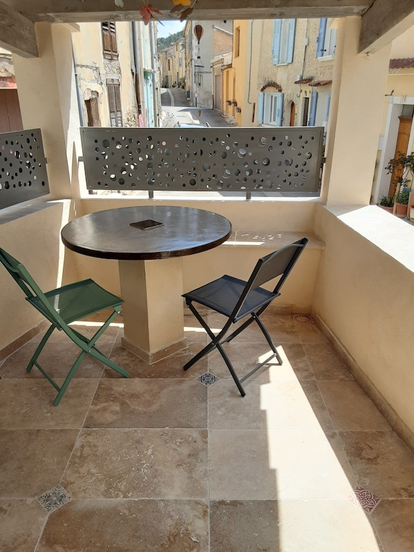 Un petit balcon avec une table ronde et deux chaises, une vue sur une rue étroite bordée d’immeubles et un paravent décoratif en métal.