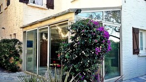 A building with a glass door entrance, a small garden with purple flowers, and a stone-tiled patio. - T3 2nd floor of the villa of the sea (Hyères)