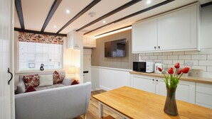 A modern kitchen with white cabinets, a wooden table, and a bench. There is a window with blinds and a sofa with decorative pillows.