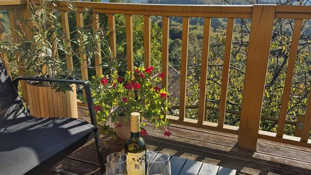 Eine Holzterrasse mit einem Tisch für zwei Personen, komplett mit einer Weinflasche, zwei Gläsern und einer Schale Trauben.