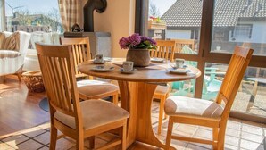 A cozy dining area with wooden chairs and a round table, a vase of flowers, and a view of the outside through large windows. - House (Simonsberg)