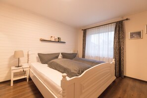 A bedroom with a white bed, a bedside table with a lamp, a shelf with books, and a window with curtains.
