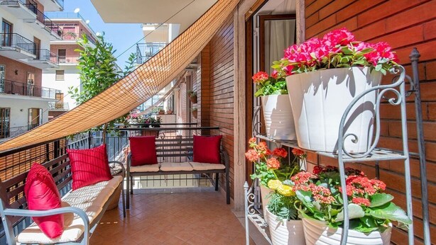 Ein Balkon mit einer hölzernen Bank, roten Kissen und einem Metallregal mit Blumentöpfen.