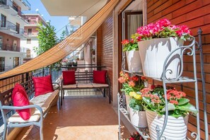 Ein Balkon mit einer hölzernen Bank, roten Kissen und einem Metallregal mit Blumentöpfen.