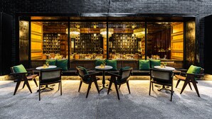 An outdoor seating area with round tables and chairs, a brick wall, and a view of the interior bar area. - Payaa Hotel (Pattaya)
