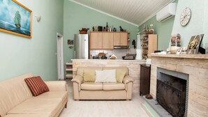 A living room with a beige sofa, a fireplace, and a kitchen area with wooden cabinets and a stone fireplace surround. - Feels Like Your Home at Ermioni (Ermionida)