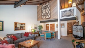 Un salón amplio con techo alto, una viga de madera en el techo y un comedor con sillas color turquesa, además de un televisor sobre un mueble de madera.