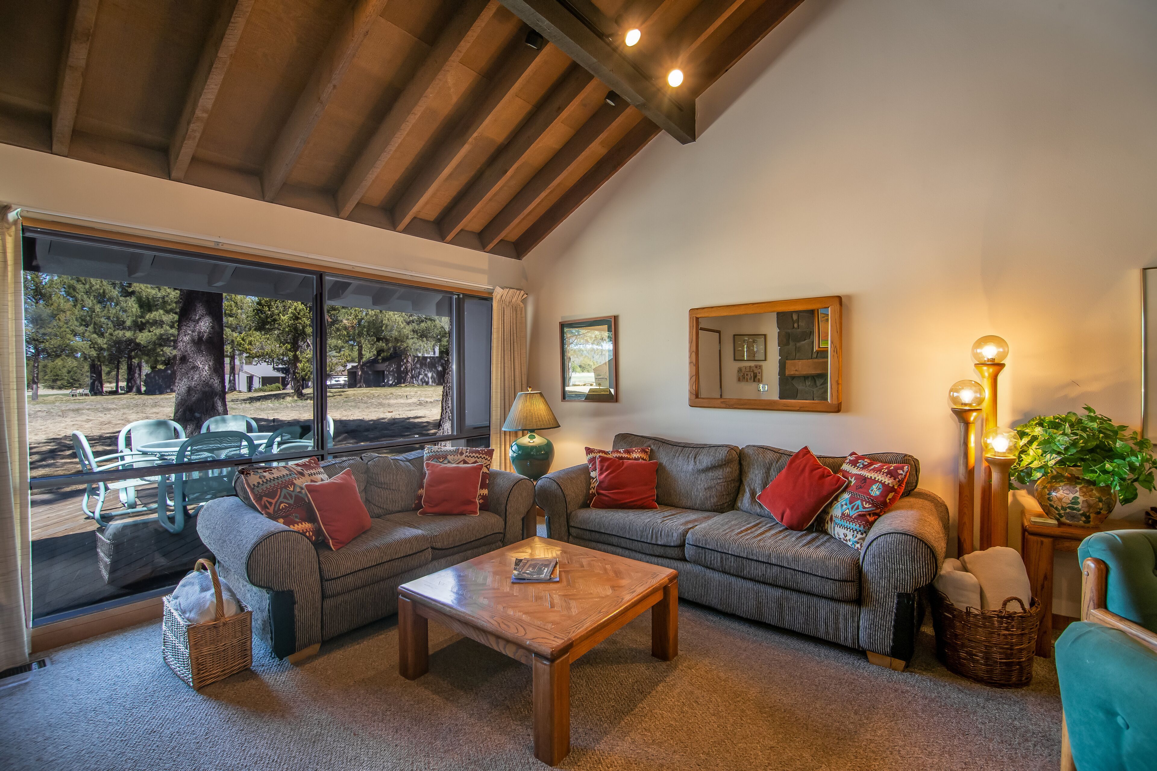A cozy living room with a wooden ceiling, a large window, a sofa set, a coffee table, and a lamp.