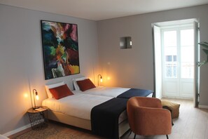A modern bedroom with two beds, a large abstract painting, a side table with a lamp, and a view of the outdoors through a door.