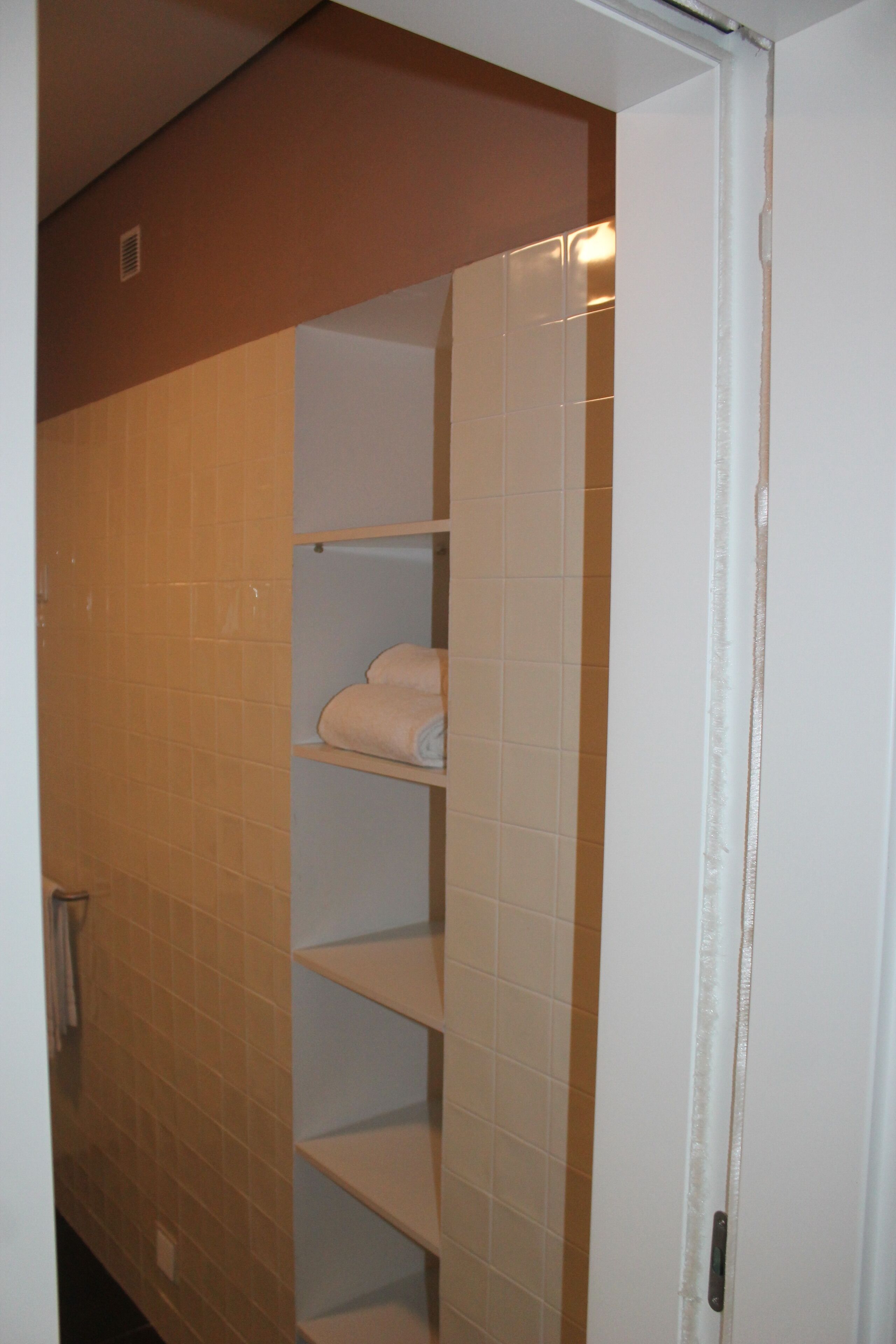 Une salle de bain avec des murs carrelés de couleur beige, une porte blanche et une étagère où sont pliées des serviettes.