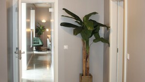 Un pasillo con suelo de madera, un taburete de ratán trenzado, una planta en maceta y una silla al fondo.