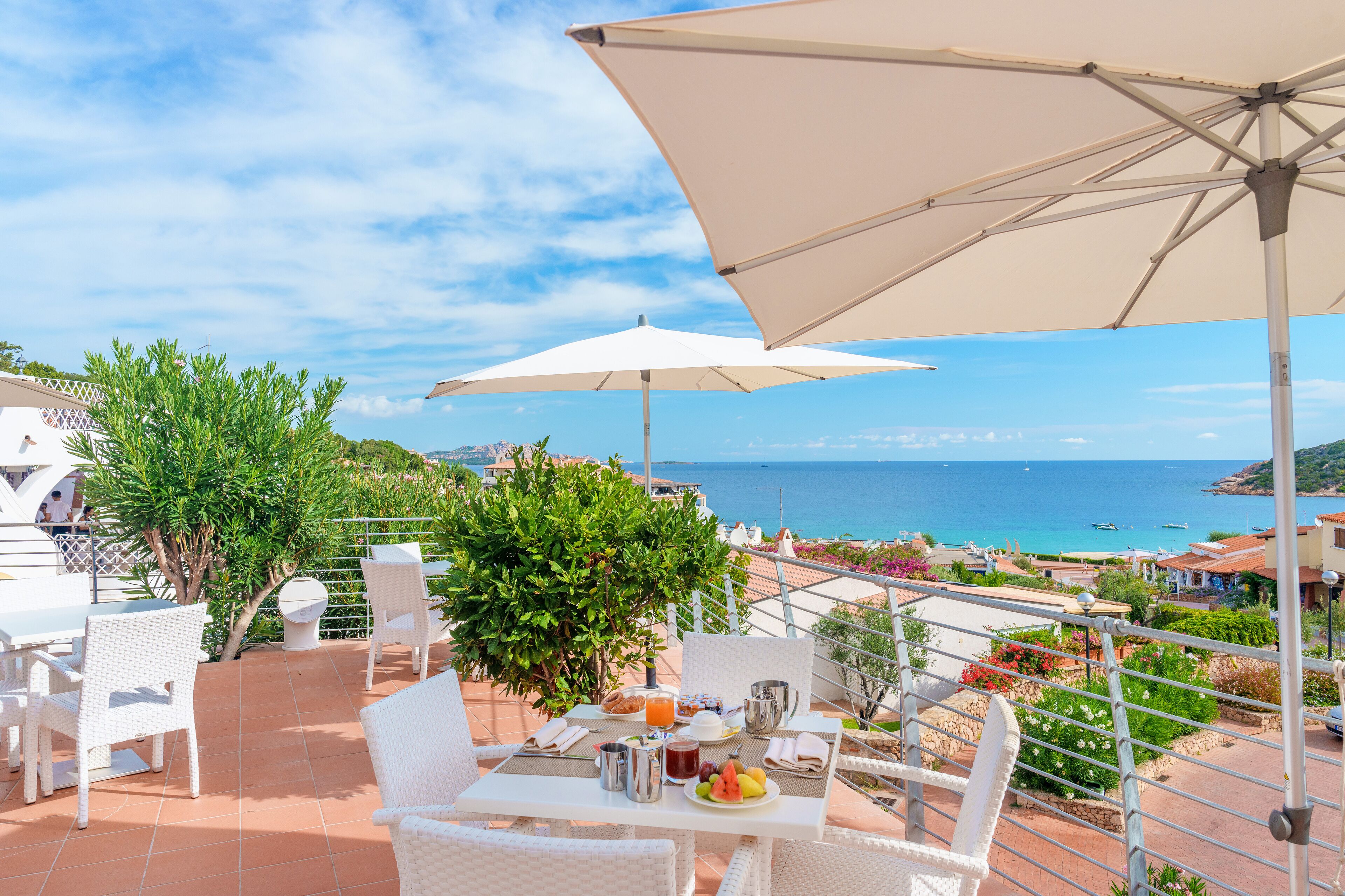 A terrace with white wicker furniture, a table set with breakfast, and a view of the sea.