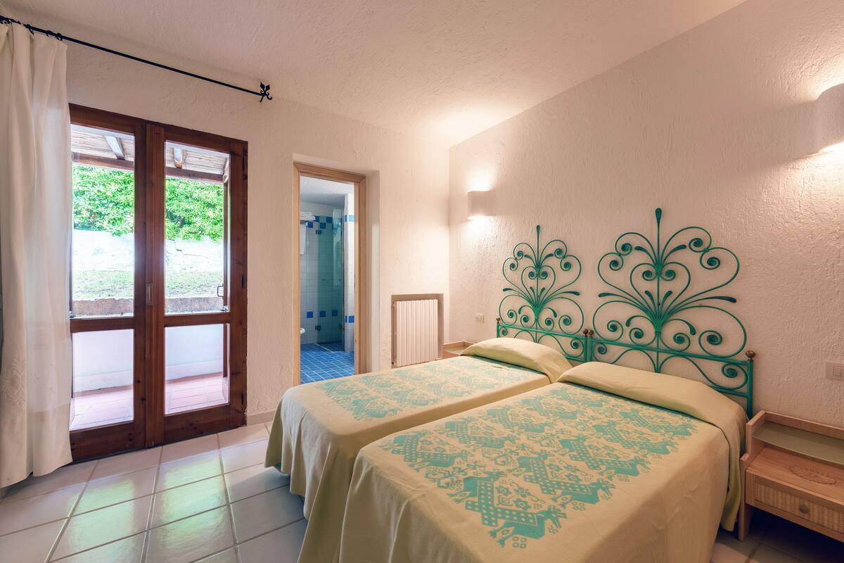 A bedroom with two beds, a wooden headboard, a nightstand, and a sliding glass door leading to a balcony.