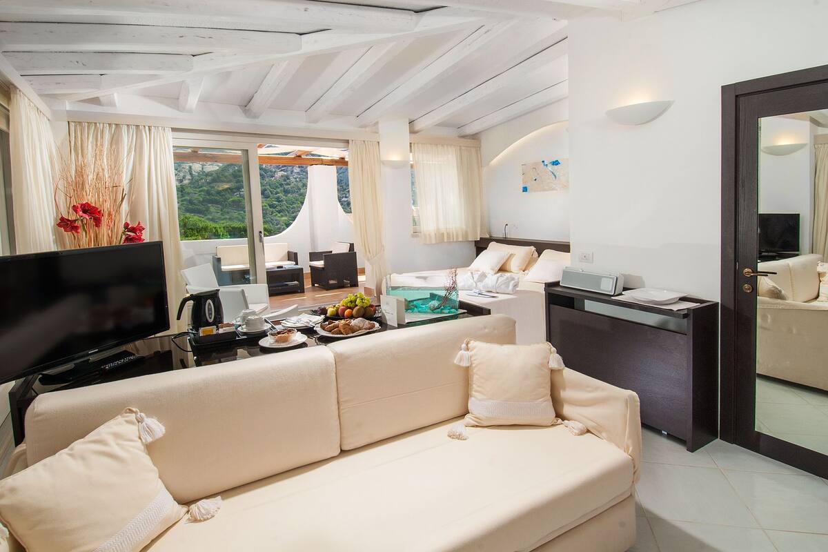 A modern living room with a sofa, a dining area with a table set for two, a TV, and a view of the outdoors.