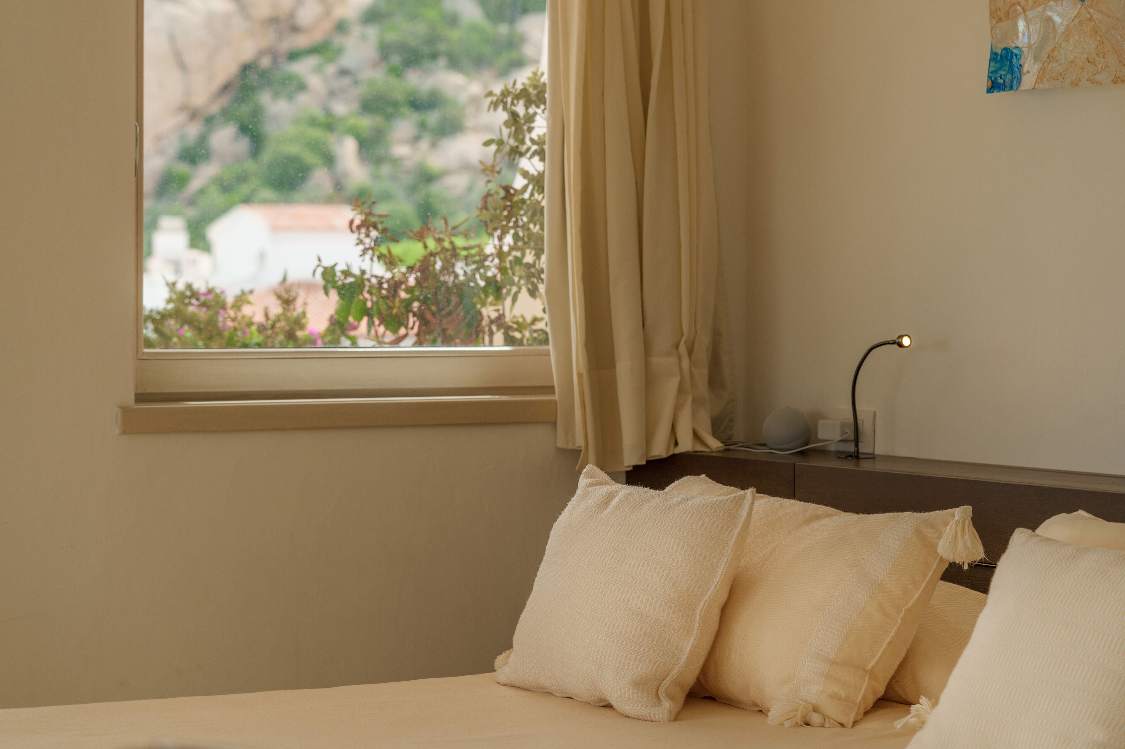 A bedroom with a bed, pillows, a nightstand, a lamp, and a window with curtains.