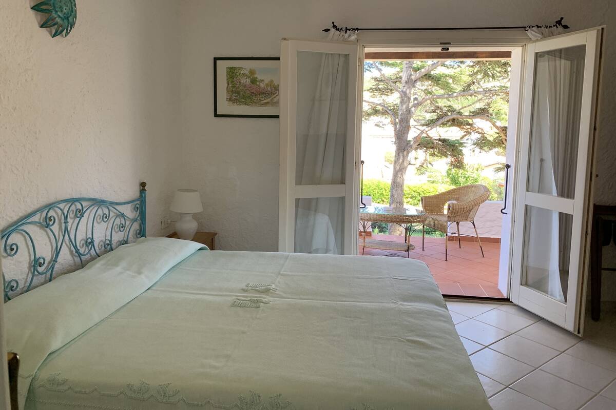 A bedroom with a bed, a nightstand, a lamp, and an open door leading to an outdoor seating area with a chair and table.