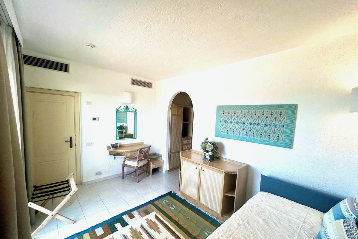 A room with a bed, a desk, a chair, a wardrobe, a mirror, and a patterned rug.