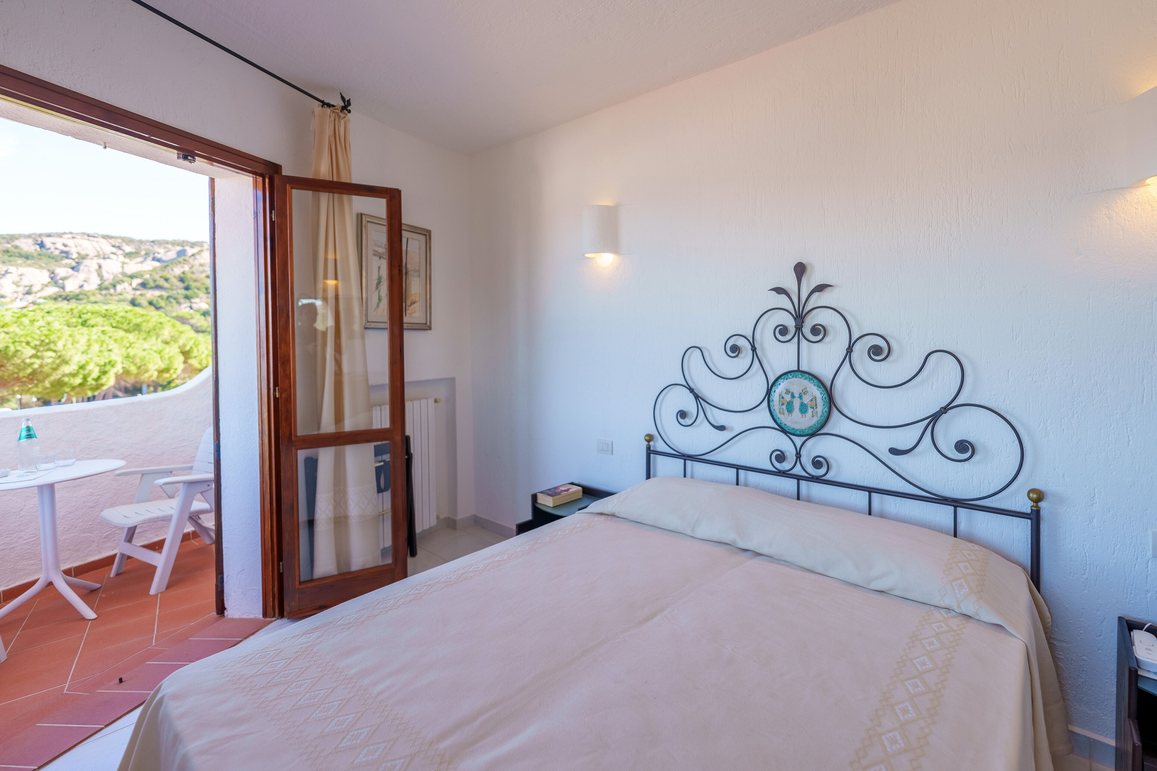 Una camera da letto con un letto, un comodino, un elemento decorativo appeso al muro e vista all'esterno attraverso una porta aperta.