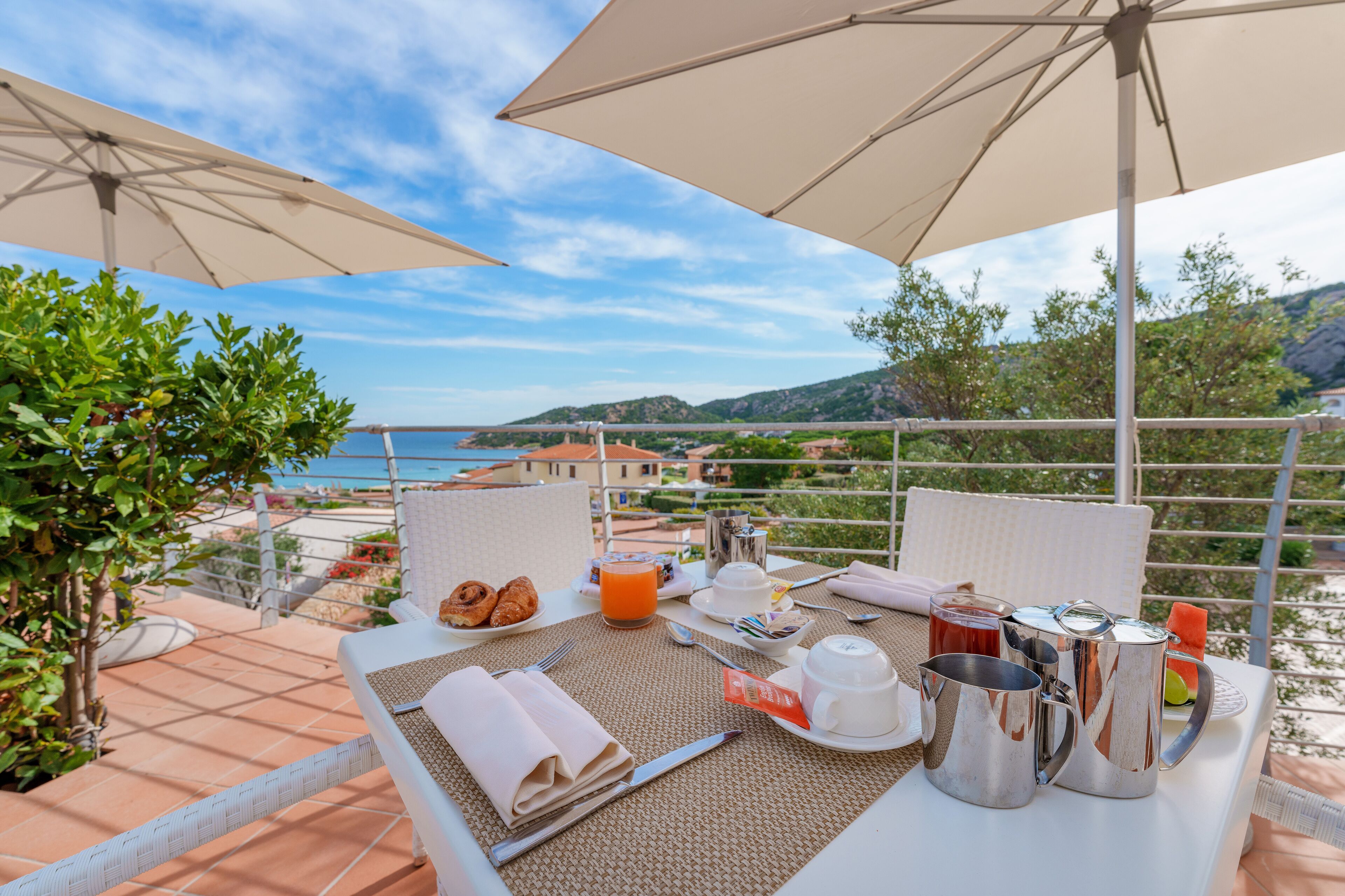 A breakfast setup on a terrace with a view of the sea, including a table with breakfast items, a view of the coastline, and outdoor seating.