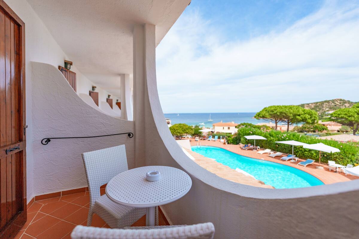 A balcony with a white chair and table, overlooking a swimming pool and a coastal view.