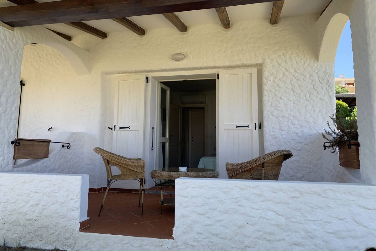 A covered patio with two wicker chairs and a table, white textured walls, and wooden beams.