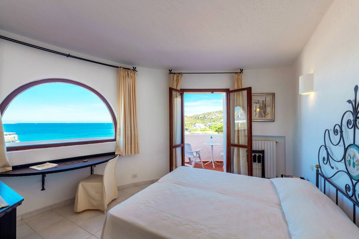 A bedroom with a large bed, a desk with a chair, a view of the sea, and a balcony with a table and chair.
