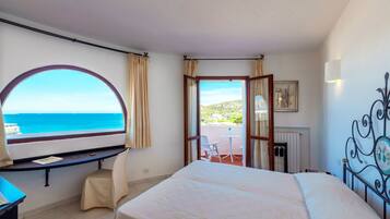 A bedroom with a large bed, a desk with a chair, a view of the sea, and a balcony with a table and chair.