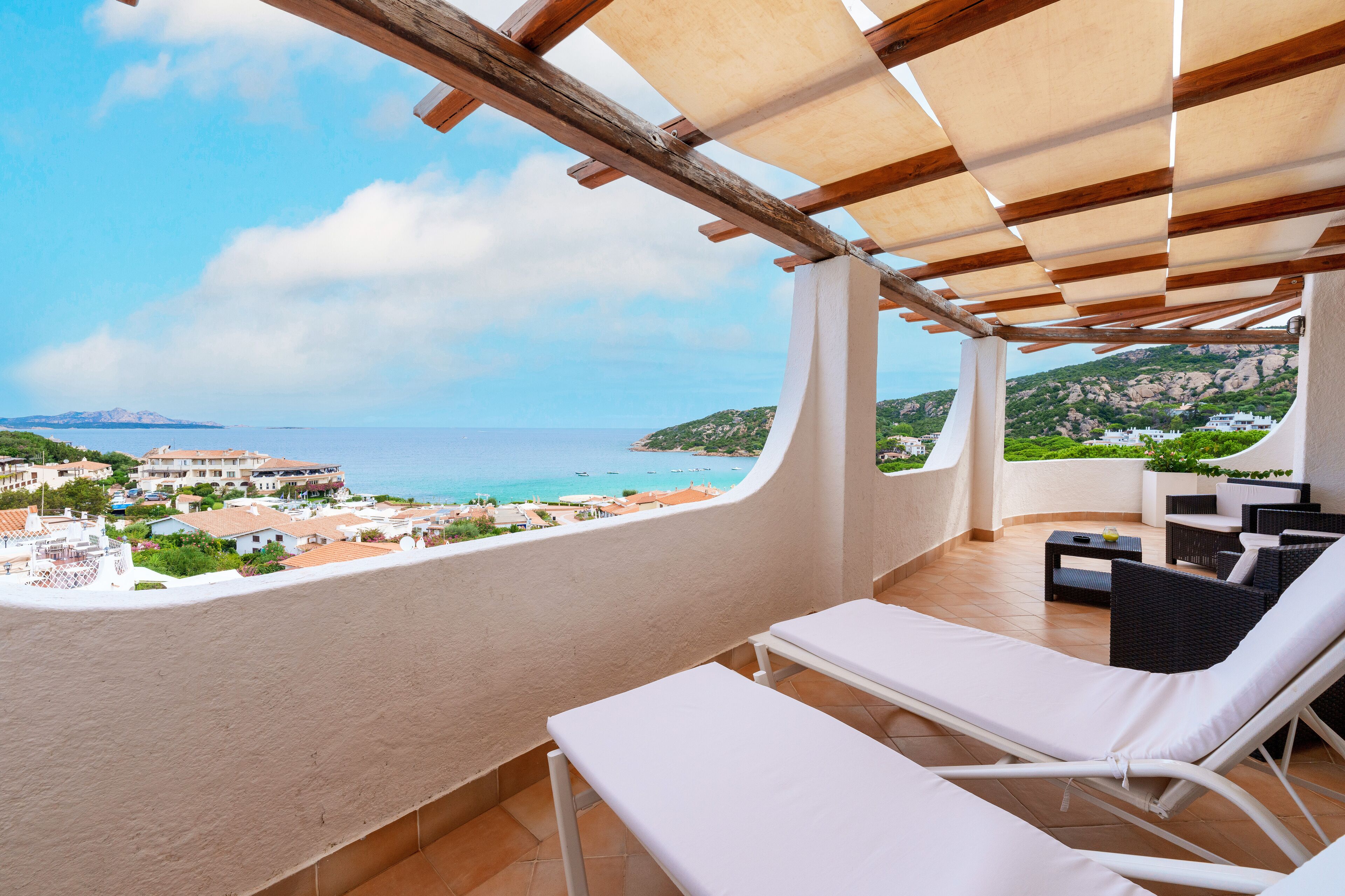 A balcony with white lounge chairs, a wooden pergola, and a view of the sea and buildings.