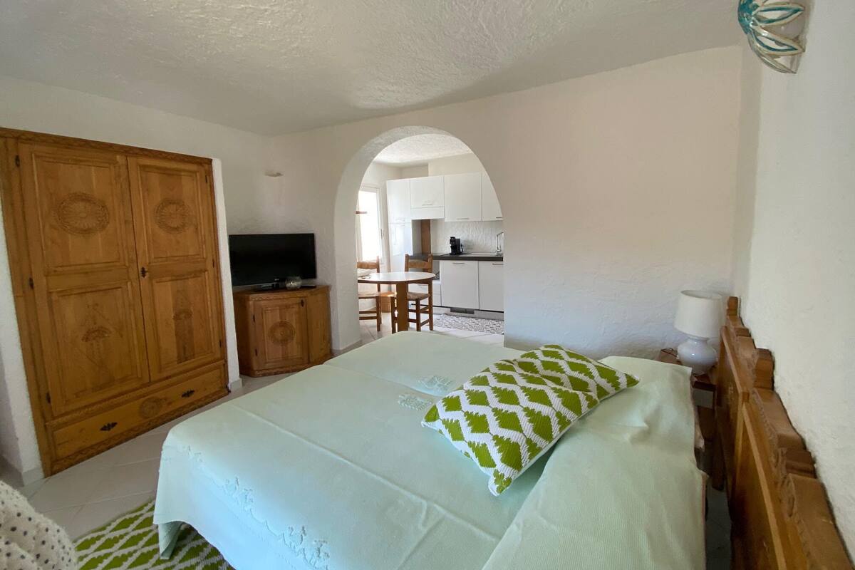A bedroom with a bed, a television on a wooden cabinet, and a kitchen in the background.